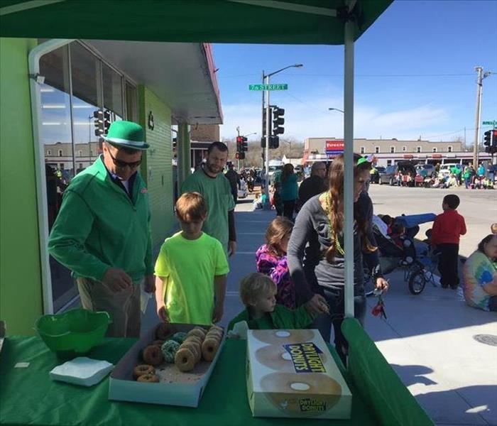 A SERVPRO employee has St Patrick's Day gear on standing in front of an outdoor tent with donuts