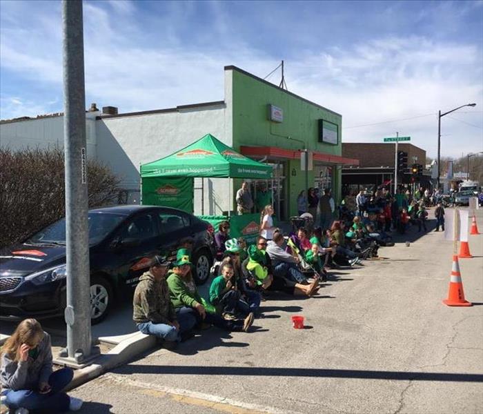 A crowd of people in St Patrick's Day gear is standing outside a SERVPRO office waiting for a parade to start